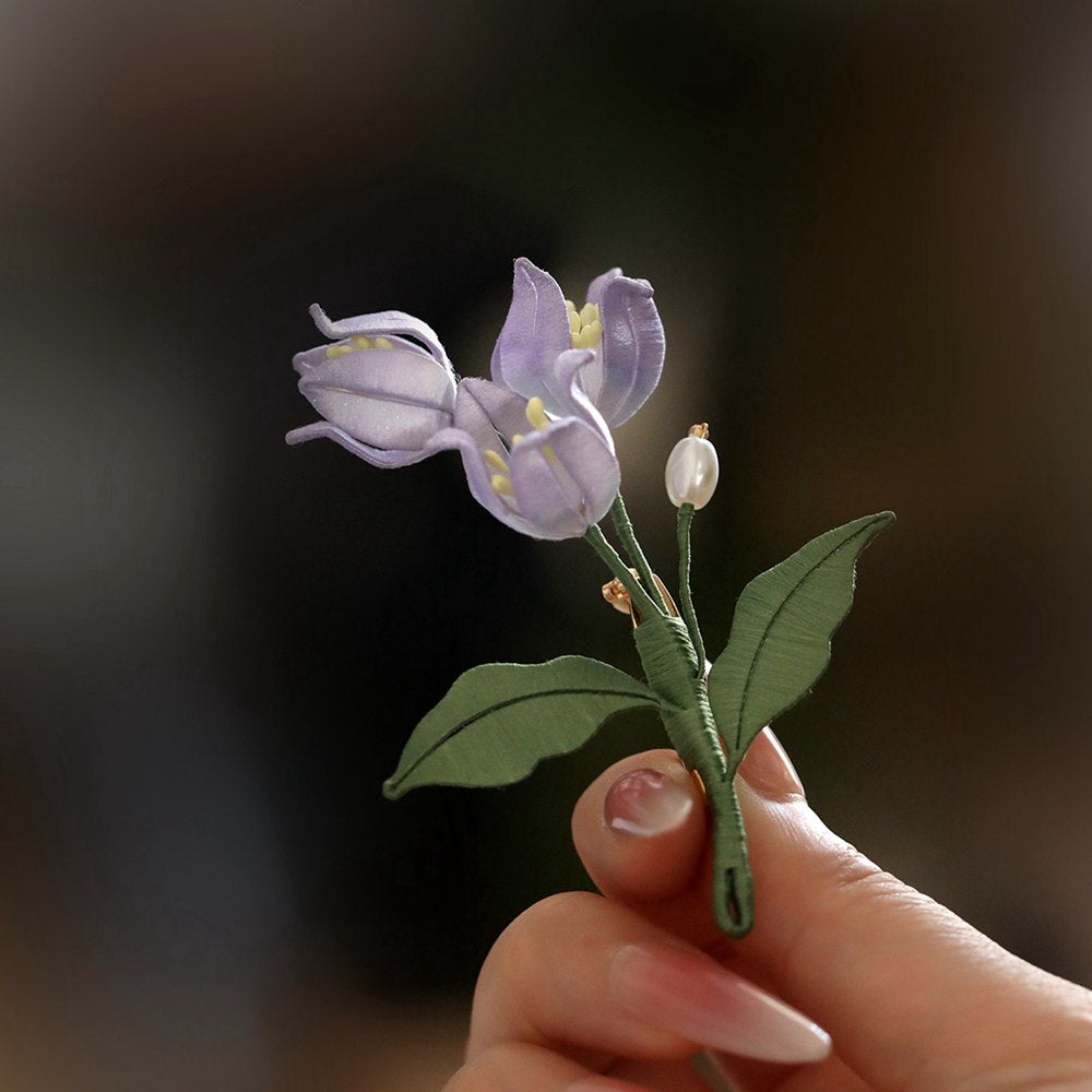 Purple brooch held in hand showing small bell-shaped blooms and pearl-like beads